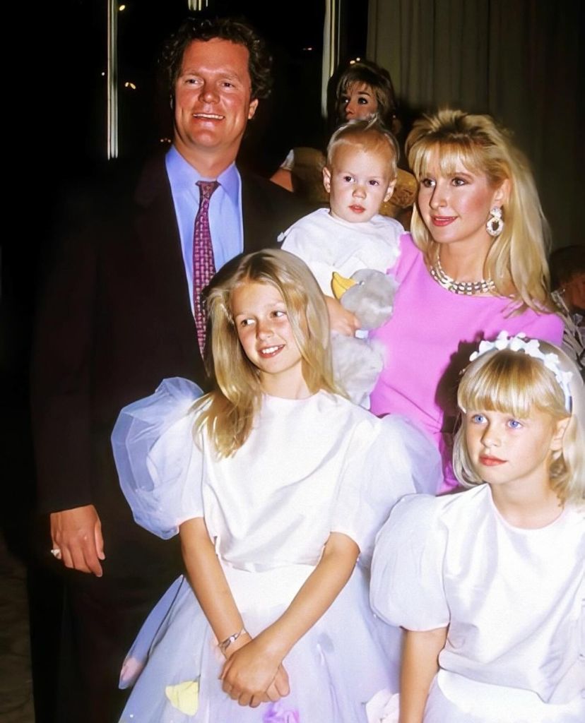 Rick Hilton stands behind his daughter Nicky, who is wearing a white bridesmaid dress, with her sister Paris standing next to her. Kathy Hilton stands holding her young son Barron in her arms, wearing a pink dress.