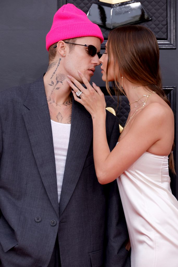 Hailey Bieber in a white dress and a pink hat is kissing her husband Justin