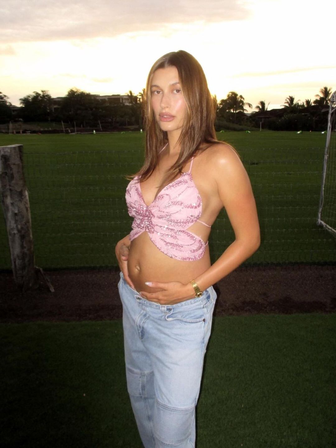 Hailey Bieber holding her baby bump while wearing a pink butterfly top