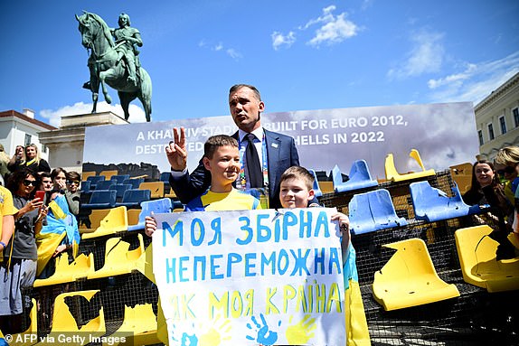 Topshot - Ukrainian Football Association president Andriy Shevchenko with children holding a poster that reads "My national team is as invincible as my country" In front of an installation of damaged seats in the colours of Ukraine taken from the Sonyachny football stadium in Kharkiv, Ukraine, in Munich, southern Germany, on June 17, 2024, where Romania vs Ukraine's UEFA Euro 2024 first round football match will take place later. The installation is dedicated to sports facilities that were bombed during the Russian-Ukrainian war. Kharkiv was one of the cities to host the UEFA Euro 2012 European Football Championship. Sonyachny Stadium was built for this competition and was later used as a training base for the Ukrainian national football team. It was destroyed by Russian shelling in May 2022. (Photo by TOBIAS SCHWARZ/AFP) (Photo by TOBIAS SCHWARZ/AFP via Getty Images)