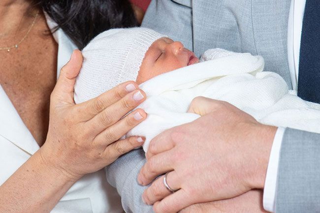 a close up photo of baby archie wrapped in a white blanket and hat with meghan and harrys hands touching him