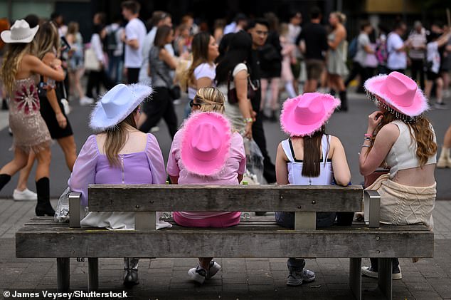 Four women wearing pink and blue cowboy hats sit down on a bench outside Wembley Stadium before the gig