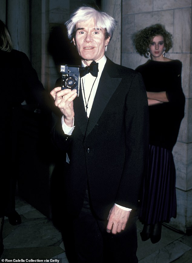 He wore a fitted, black suit jacket that buttoned down the front, along with a pair of loose-fitting black trousers; Warhol seen in 1985