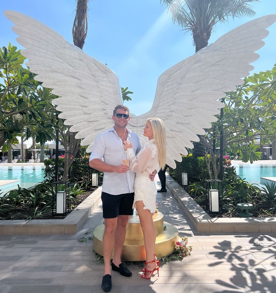Amelia Spencer and Greg Mallett in front of angel wings by the pool 