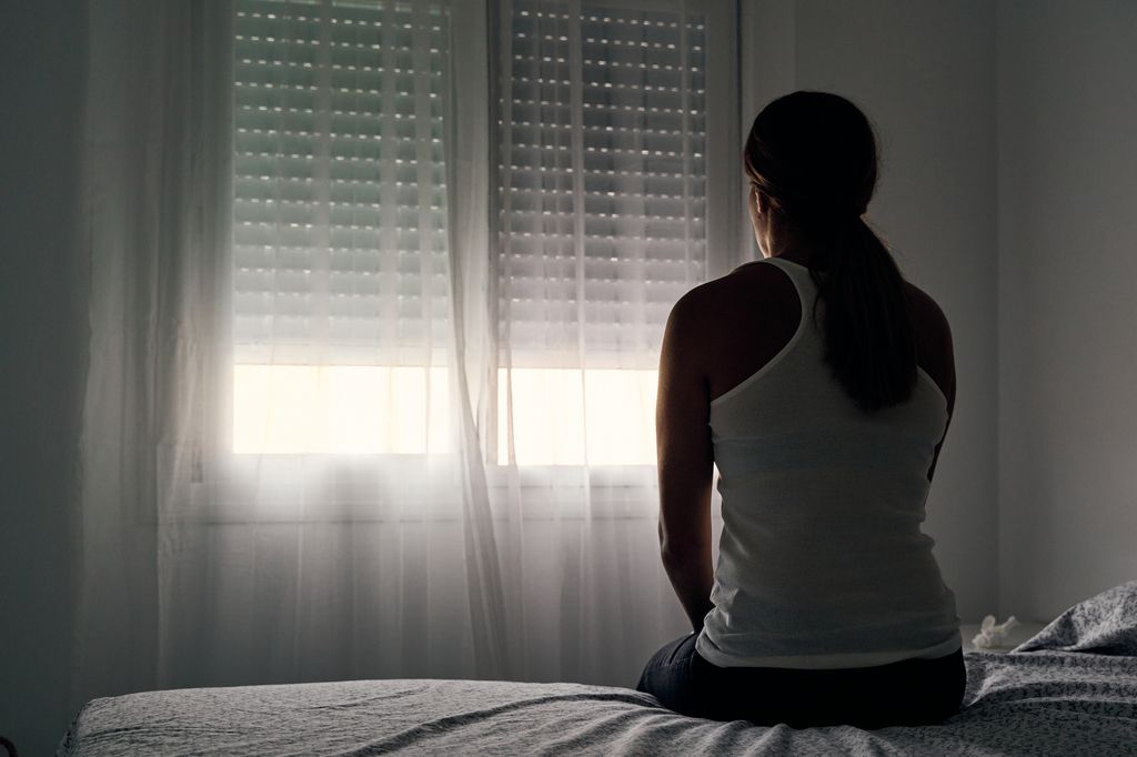 In the background, an unidentified woman sits on her bed, looking out the window.