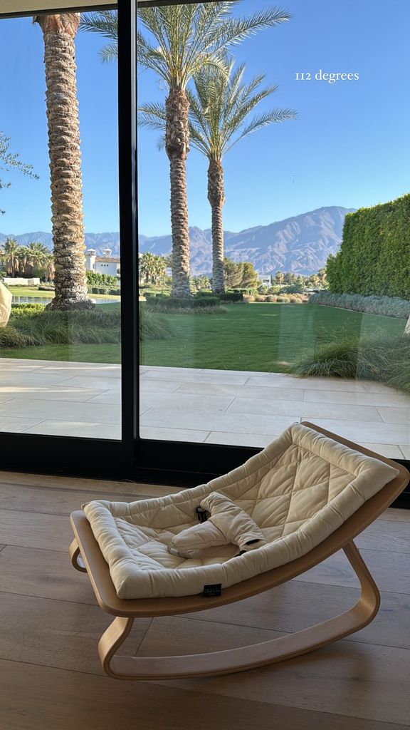 Photo posted by Kourtney Kardashian. Inside is a white rocker chair placed on the wooden floor, and outside are palm trees on the floor-to-ceiling glass windows